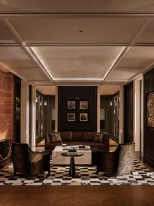 Luxurious hotel lobby lounge with two zebra-print velvet armchairs, a brown leather sofa, white marble coffee table, and black-and-white checkerboard cowhide rug under dramatic coffered ceiling with cove lighting.