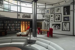 Modern double-height living room with curved light-grey sectional sofas, a large gallery wall of black and white portrait photography, red velvet dining chairs, wooden table, and industrial metal structural columns under a skylight.
