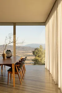 Modern dining room featuring a long walnut table, black leather dining chairs, large floor-to-ceiling windows overlooking mountains and fields, with sheer white curtains and minimalist decor.
