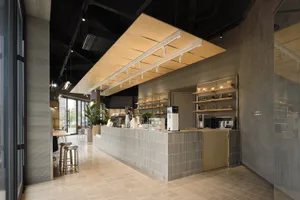 Modern minimalist cafe interior featuring a long grey concrete tile service counter, wooden canopy lighting fixture, metal bar stools, and open shelving against concrete walls.