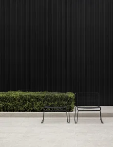 Two black metal wire chairs sitting on a light grey tiled patio in front of a dark corrugated wall and a row of green bushes.