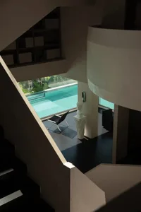 View from above of a modern curved concrete staircase overlooking a contemporary living area with a white classical bust on a pedestal, black leather chair, and large glass windows revealing an infinity pool surrounded by tropical greenery.