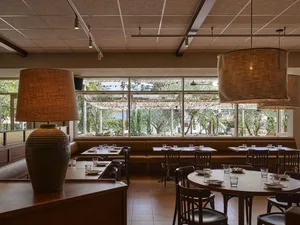 Warmly lit restaurant interior featuring round and rectangular wooden tables, dark wood chairs, built-in leather banquettes, large woven pendant lamps, and a prominent terracotta table lamp in the foreground.