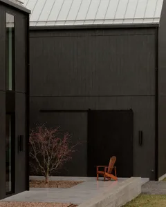 Modern black charred wood house exterior with concrete patio, small red-leaf tree, and orange Adirondack chair in a minimalist courtyard.