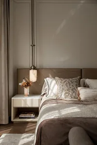 Modern luxury bedroom featuring a taupe leather upholstered headboard, marble pendant lamp, neutral bedding with floral accent pillows, and a white marble-top nightstand with dried floral arrangement.