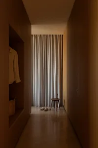 Narrow walk-in closet featuring floor-to-ceiling walnut wood cabinetry, a small wooden stool, white linen curtain, and woven basket storage.