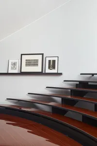 Modern curved walnut wooden staircase with glossy finish, black floating shelves displaying framed black-and-white photographs against a white wall.