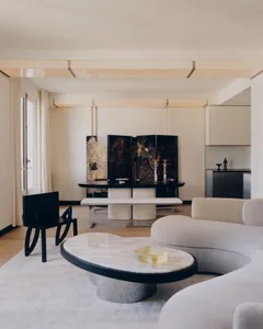 Modern minimalist living and dining area featuring a curved white marble coffee table, cream bouclé sectional sofa, black sculptural chair, and a large four-panel black lacquered folding screen with gold artwork.
