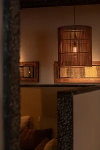 Warm interior view of a living room featuring a large cylindrical woven bamboo pendant lamp, textured stone wall elements, wooden framed textile art, and neutral-toned cushions in earthy brown and beige.
