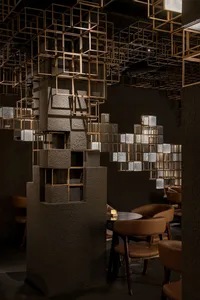 Interior of a stylish restaurant featuring textured concrete columns, brown leather dining chairs, round tables, and an elaborate ceiling installation of illuminated golden cubic metal frames.