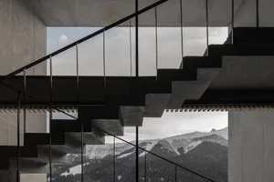 Modern concrete staircase with black metal railings overlooking snow-capped mountains and forested valleys.