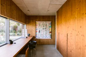 Modern home office interior featuring vertical wood paneling, a long wooden desk, black leather chair, large colorful abstract painting, and concrete ceiling and floor.