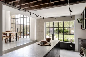 Modern industrial-style kitchen with polished concrete island, dark wood ceiling beams, black steel-framed windows overlooking a garden, and wooden dining table in the background.