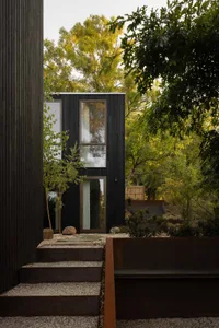 Modern dark charred wood cabin exterior with large windows, surrounded by green trees and accessed via concrete and gravel steps in a forested setting.
