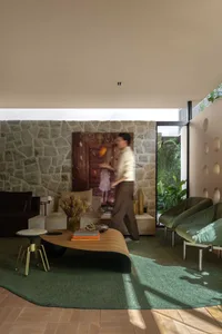 Modern living room featuring a large stone accent wall, curved wooden coffee table, green velvet lounge chairs, and a view to an outdoor garden with a person walking through the space.