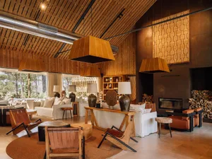 Spacious modern rustic living room featuring a high wood-paneled cathedral ceiling, large leather sling chairs, white linen sofas, a concrete fireplace with stacked firewood, and multiple large woven leather pendant lights.