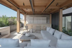 Modern rooftop terrace with light grey outdoor sofa, round travertine coffee table, built-in outdoor kitchen in travertine, wooden ceiling beams and abundant greenery under a clear blue sky.
