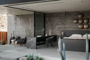 Modern minimalist bedroom and home office with raw concrete walls, black marble desk, dark wood lounge chairs, and desert cacti plants.