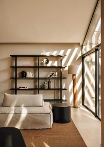 Minimalist living room corner featuring a black open shelving unit displaying ceramics and books, a cream linen daybed, round black side table, tall oak floor lamp, and large glass doors letting in strong natural light that casts striped shadows on the wall and floor.