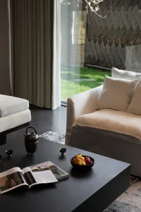 Modern minimalist living room featuring a cream sofa, dark wood coffee table with tea set and magazine, and a striking dark wood chevron accent wall beside a large glass door opening to a green garden.