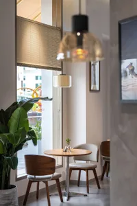 Modern minimalist cafe interior featuring a round wooden table, walnut and upholstered dining chairs, large potted plant, and warm pendant lights by a window with blinds.