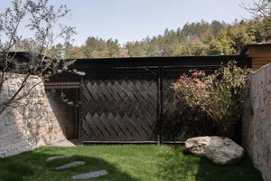 Modern garden featuring a large chevron-patterned black bamboo fence between rustic stone walls, with stepping stones on a green lawn and a large natural boulder in the foreground.