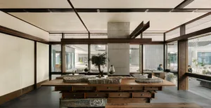 Spacious modern design studio interior featuring a large reclaimed wood worktable with stone models, a central concrete structural pillar, dark metal beams, and large glass windows overlooking a garden courtyard.