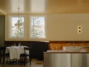 Elegant restaurant interior featuring dark walnut wainscoting, a bright orange upholstered banquette, round tables with white linens, dark wood chairs, and two large windows overlooking pine trees and the ocean.