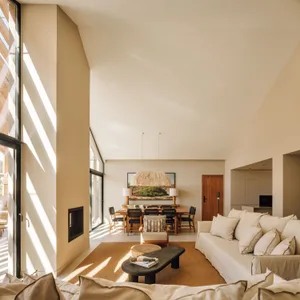 Bright, airy open-plan living room with high ceilings, large windows, beige linen sofa, wooden dining table, woven pendant light, and jute rug bathed in strong natural sunlight.