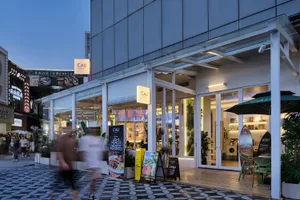 Modern cafe named CALI with large glass windows, white structural frames, outdoor seating, and potted plants along a patterned stone pedestrian street at dusk.
