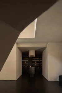 Modern minimalist butler pantry viewed through a dramatic angular concrete opening, featuring dark micro-cement walls, black fluted island, and illuminated open shelving.