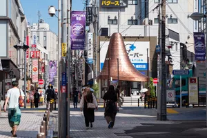 Pedestrians walking on a sunny day along a Japanese shopping street featuring a large copper cone-shaped public art installation in the background.