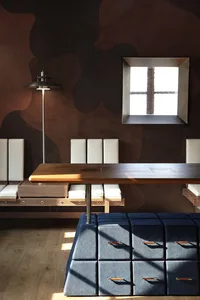 Modern dining room featuring a long walnut table, built-in white banquette, navy modular ottomans, a sculptural floor lamp, and a large abstract brown organic mural wall with a square window.