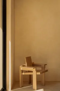 A solid oak armchair with blocky geometric form sits in the corner of a minimalist room with warm beige plaster walls and concrete floor, illuminated by natural sunlight from the side.