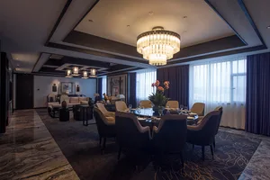 Luxurious open-plan dining and living area featuring dark marble flooring, a large round dining table with upholstered chairs, and an oversized crystal chandelier hanging from a coffered ceiling.