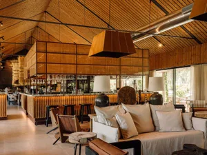 Modern rustic bar lounge featuring a soaring vaulted wooden ceiling, a long reclaimed wood bar with rattan screens, cream linen sofas with plush pillows, and large pendant lights in a spacious, airy interior.