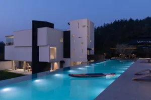 Modern white concrete villa with black architectural accents and a long illuminated infinity pool at dusk, with lounge chairs and forested hillside in the background.