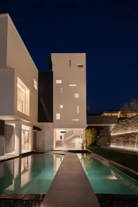 Night view of a modern concrete villa with a long rectangular reflecting pool, illuminated windows, and a central courtyard under a deep blue night sky.