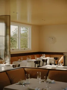Elegant restaurant interior with warm yellow walls, terracotta leather banquette seating, white-clothed dining tables, dark wood chairs, and large windows overlooking greenery.