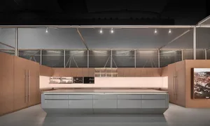 Modern minimalist kitchen interior with light oak wood cabinetry, a large grey central island, polished concrete floor, and dramatic linear track lighting against a backdrop of large windows with blinds.