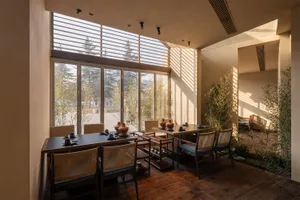 A modern Japanese-style dining room featuring dark wood tables, woven rattan chairs with blue cushions, large glass windows with wooden shutters overlooking bamboo and trees, and potted bamboo plants inside.