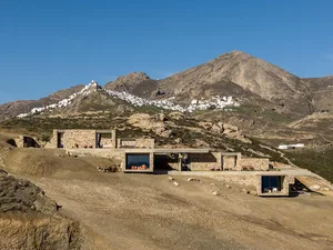 Modern stone villa with large glass windows and outdoor terraces built into a dry hillside overlooking a white Cycladic village on a mountain in Greece