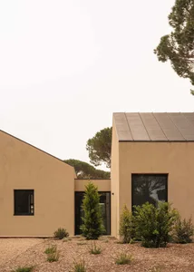 Exterior view of a minimalist modern house with beige stucco walls, black-framed windows, flat roof, and drought-tolerant garden with gravel and native shrubs under a bright sky.