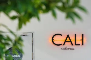 Orange neon sign with the word CALI mounted on a white wall, with green plant leaves in the foreground and a glass door with a circular kitchen sign visible to the left.