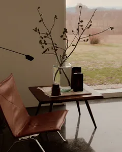 Cognac leather butterfly chair beside a walnut side table holding a large glass vase with budding branches, dark wooden objects, and a view of a winter landscape through a large window.