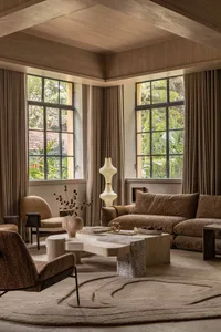 Modern organic living room featuring a large L-shaped brown bouclé sofa, sculptural travertine coffee table, boucle armchair, wooden ceiling beams, and tall windows overlooking lush greenery.