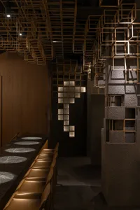 Modern restaurant bar interior featuring an intricate brass cube grid ceiling installation, textured concrete walls, and a long dark bar counter with backlit circular stone inlays and leather stools.