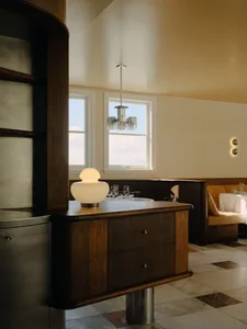 Interior view of a sophisticated restaurant featuring a curved walnut sideboard with a glowing spherical lamp, set against a dining area with banquette seating and large windows.