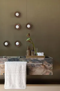 Modern bathroom featuring a floating vanity made of multicolored marble with an olive-green wall, wooden wall sconces, and a white towel hanging on a metal rod.