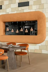 Modern restaurant dining area featuring a large organic-shaped terracotta plaster pass-through window, dark wood table, burnt orange velvet chairs, and textured beige tile wall.